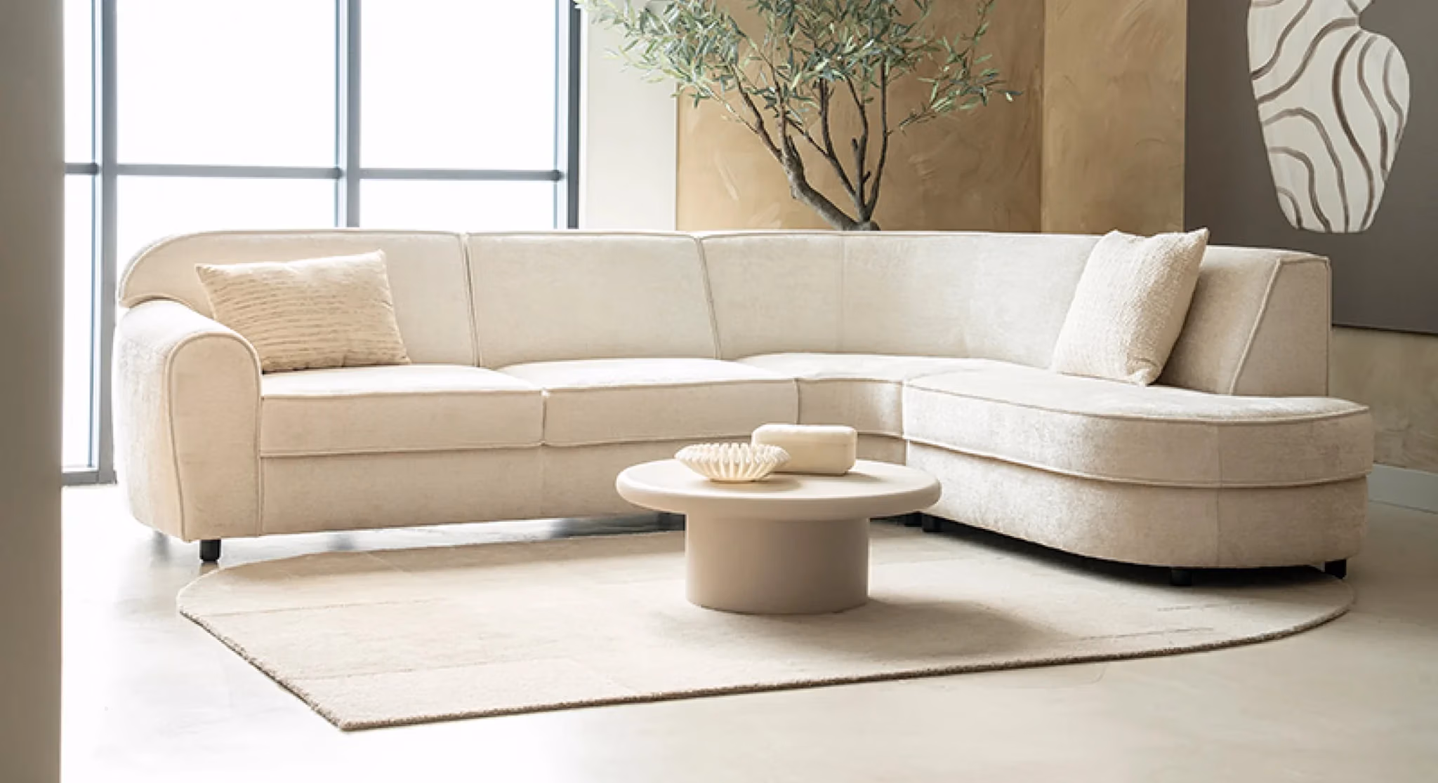 Een moderne woonkamer met een crèmekleurige hoekbank van Sevn zitmeubelen, twee kussens met structuur, een ronde salontafel met decoratieve spullen, een beige vloerkleed, een groot raam, een boom in een pot en abstracte wandkunst.