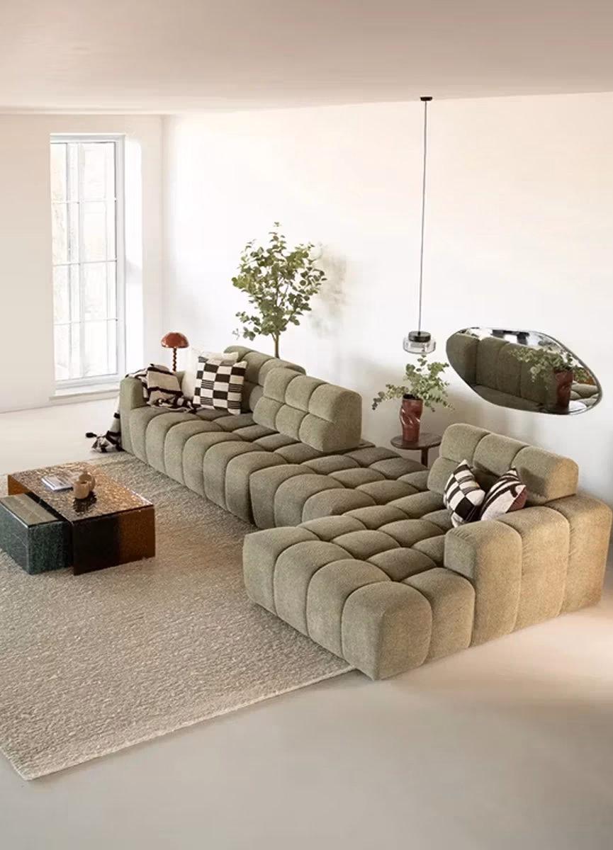 Een moderne woonkamer met een groene modulaire bank van Sevn zitmeubelen, geometrische kussens, twee vierkante salontafels, een groot raam, een vloerkleed, een kamerplant, een hangende spiegel en een minimalistische inrichting.