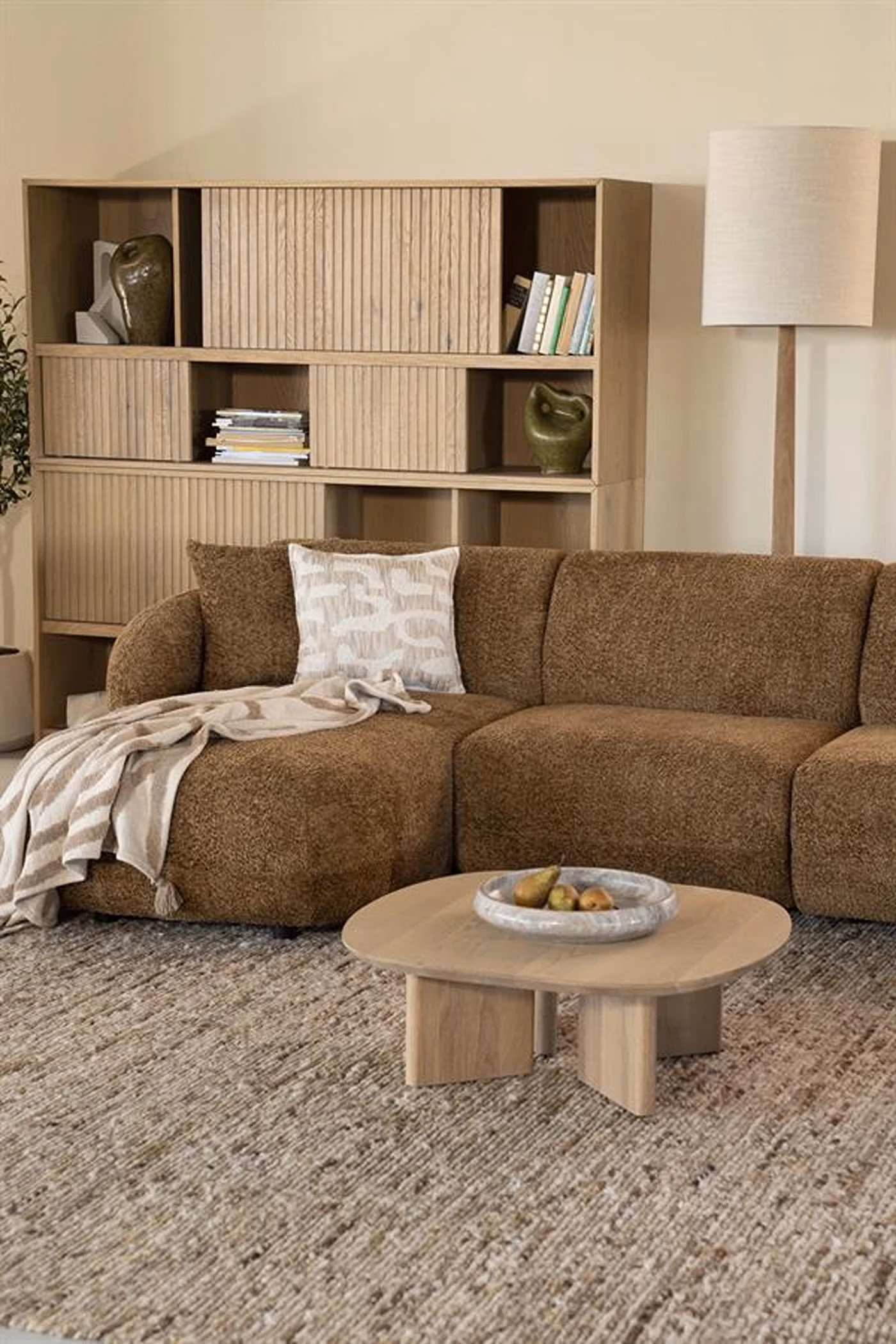 Gezellige woonkamer met een bruine Sevn zitmeubelen-hoekbank, een kussen met patroon, een lichte sjaal, een ronde houten salontafel, een groot tapijt met textuur, een houten boekenkast met boeken en decoratie, en een hoge staande lamp.