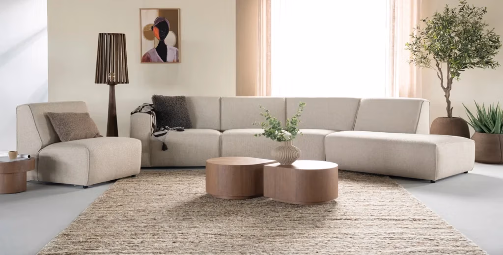 Moderne woonkamer met een beige hoekbank van Sevn zitmeubelen, twee ronde houten salontafels, een staande lamp, potplanten, een tapijt met structuur, vitrage en abstracte wandkunst aan een lichtgekleurde muur.