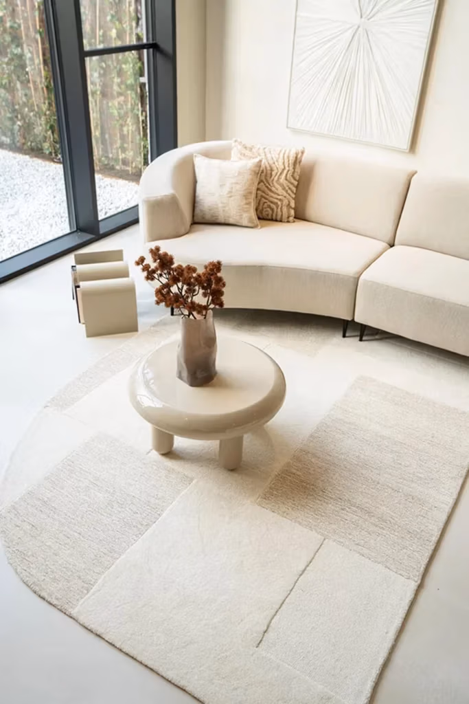 Minimalistische woonkamer met een gebogen crèmekleurige bank van Sevn zitmeubelen, een kussen met patroon, abstracte wandkunst, een ronde salontafel met een vaas, boeken en een wit vloerkleed met textuur bij de grote ramen.