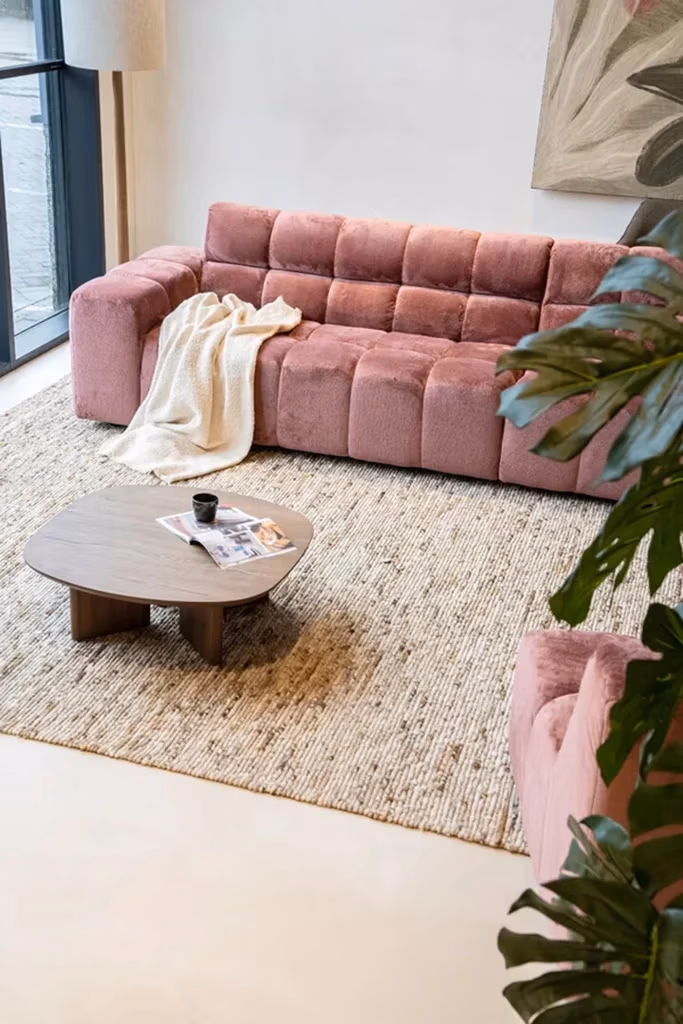 Een moderne woonkamer met zitmeubelen van Sevn, waaronder een roze gecapitonneerde bank en een bijpassende fauteuil, aangevuld met een beige tapijt met structuur, een houten salontafel met daarop een kopje en tijdschriften, een lichte sjaal, een grote plant en een raam dat natuurlijk licht binnenlaat.