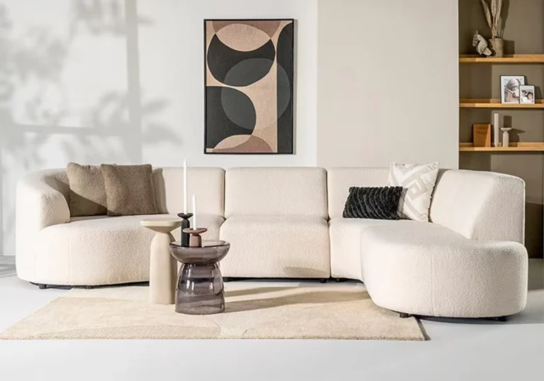 Een moderne woonkamer met een crèmekleurige hoekbank van Sevn zitmeubelen, diverse sierkussens, een licht tapijt, twee minimalistische salontafels, abstracte wandkunst en open rekken met decoratieve voorwerpen in een ruimte met neutrale tinten.