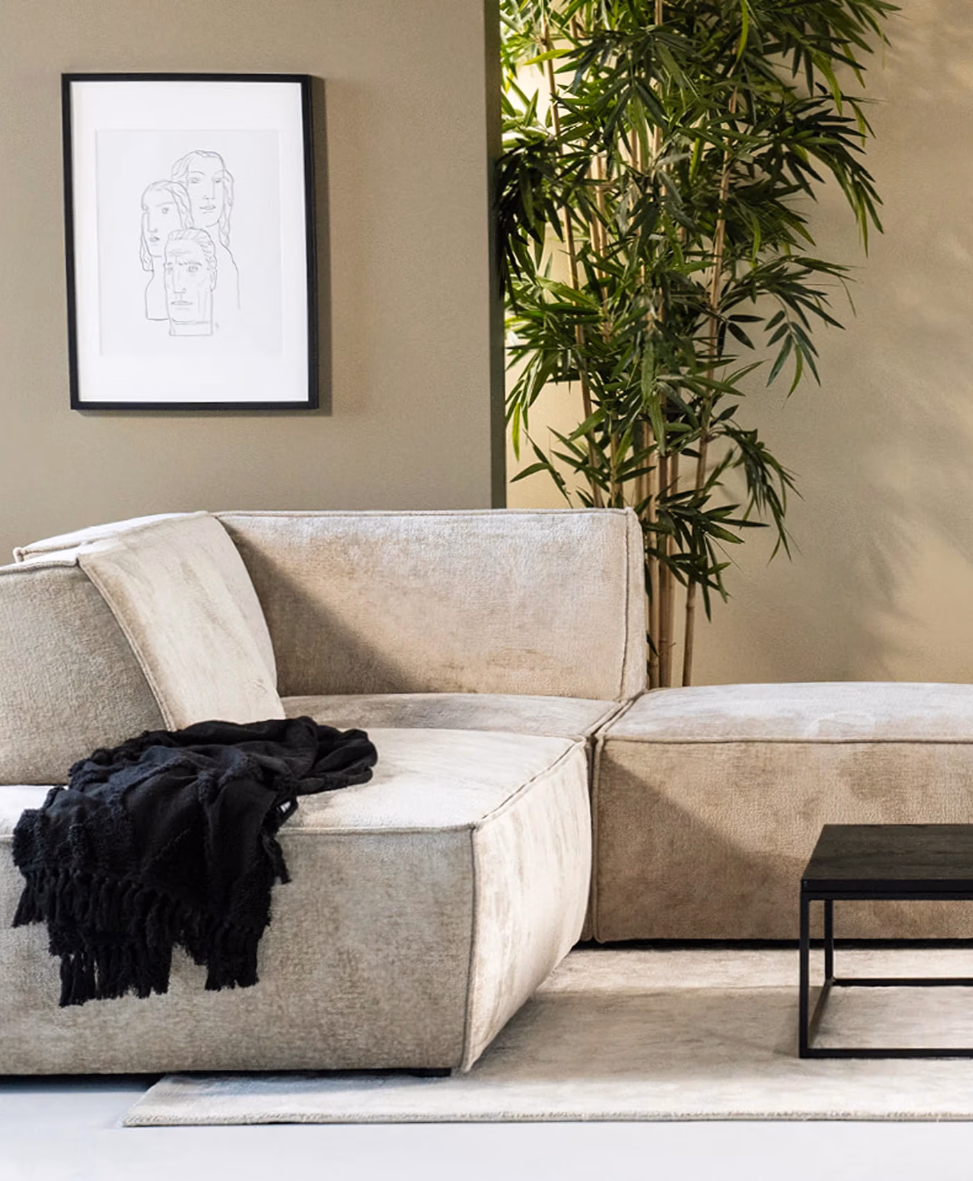 Een moderne woonkamer met een beige Sevn zitmeubelen-hoekbank, een zwarte plaid, een zwarte salontafel, ingelijste lijntekeningen aan de muur, een hoge groene plant en neutraal gekleurde muren en vloerkleed.