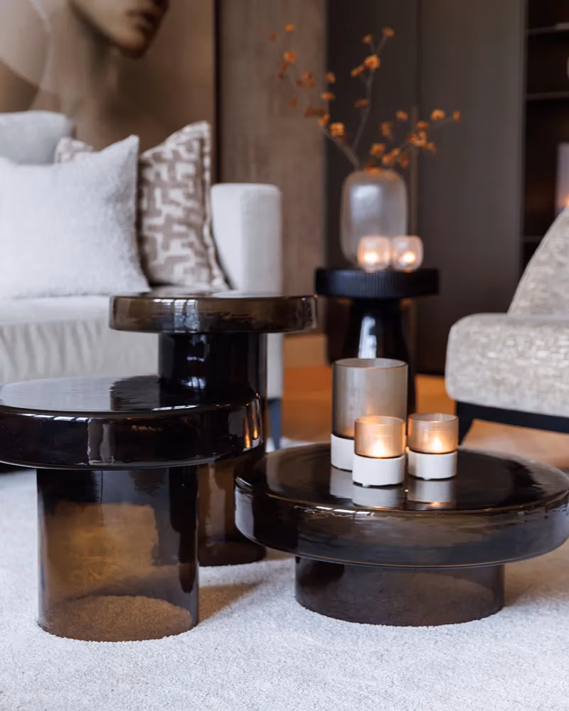 Moderne woonkamer met glanzende, donkere ronde salontafels, drie brandende kaarsen in glazen houders, een lichte bank, een kussen met patroon en op de achtergrond een vaas met takken. De neutrale en gezellige inrichting is perfect voor iedereen die zijn woonkamer op een moeiteloze manier in stijl wil vernieuwen.