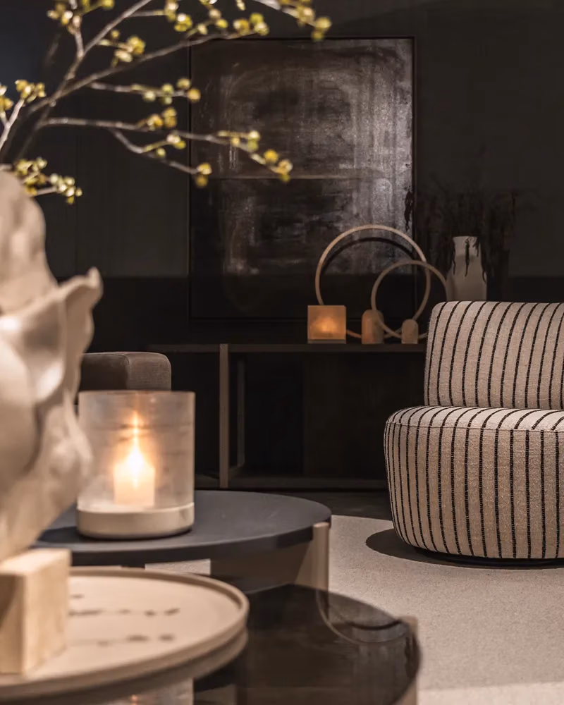 Een gezellige woonkamer met een gestreepte fauteuil, een ronde zwarte tafel, kaarsen, abstracte decoraties en een schilderij in een donkere lijst aan de muur, zacht verlicht voor een warme, uitnodigende sfeer – de perfecte inspiratiebron als je jouw woonkamer wilt vernieuwen.