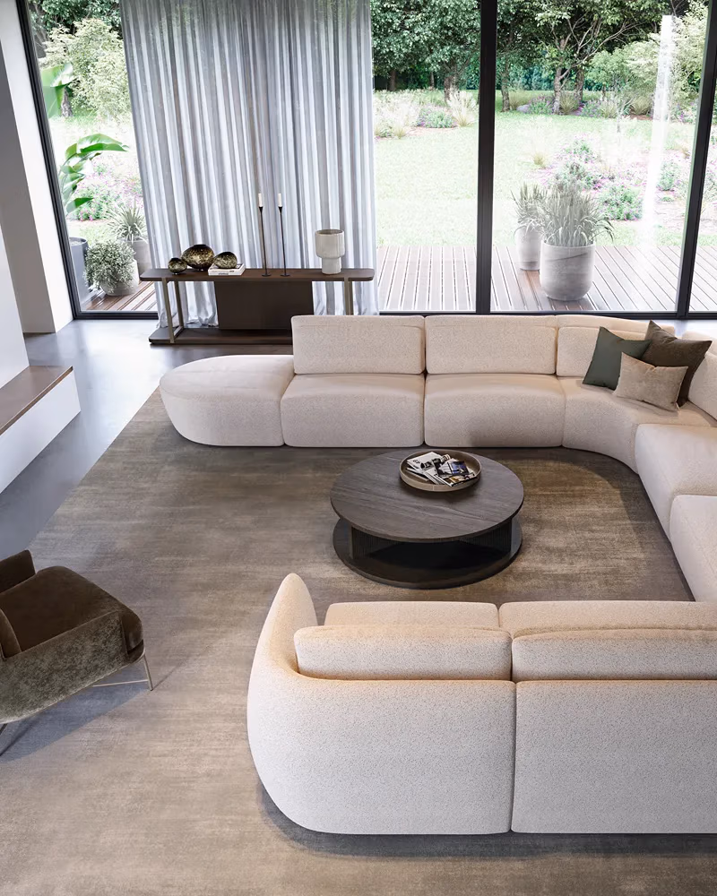 Moderne woonkamer met een grote crèmekleurige hoekbank, een ronde salontafel, een groene fauteuil, een neutraal vloerkleed en kamerhoge ramen die uitkijken op een tuin met planten en een terras – de perfecte inspiratiebron als je je woonkamer wilt vernieuwen.