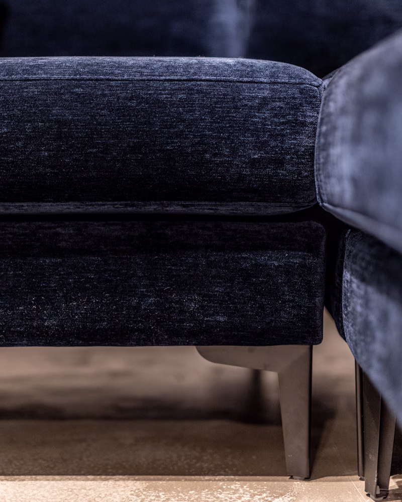 Close-up van een donkerblauwe fluwelen bank met houten poten, waarop de textuur van de bekleding en een deel van het zitkussen te zien zijn. De nadruk ligt op de onderrand en de onderkant van het meubelstuk – perfecte inspiratie als je bezig bent met het uitzoeken van een bank voor je woonkamer.