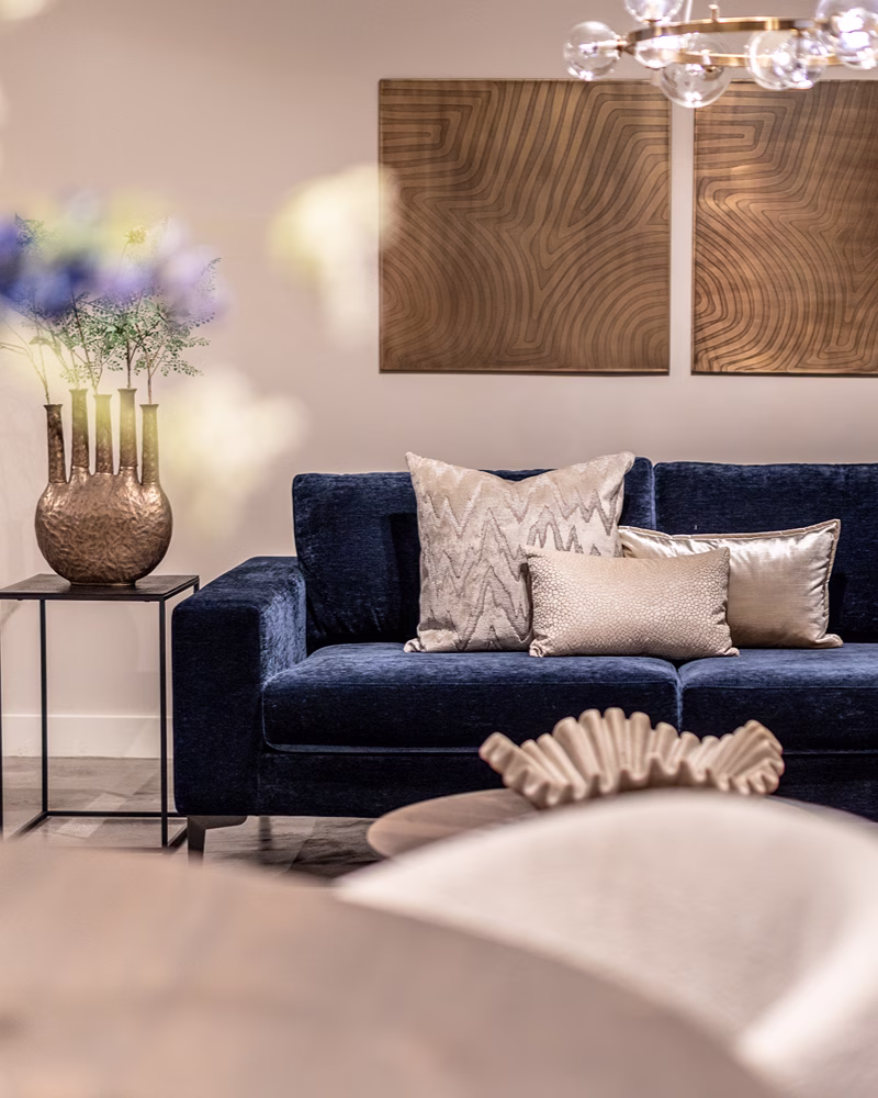Een moderne woonkamer met een donkerblauwe fluwelen bank – zorgvuldig uitgekozen om bij het interieur te passen –, kussens met neutrale patronen, een metallic vaas met bloemen op een bijzettafeltje, abstracte wandkunst en zachte verlichting zorgen voor een gezellige, elegante sfeer.
