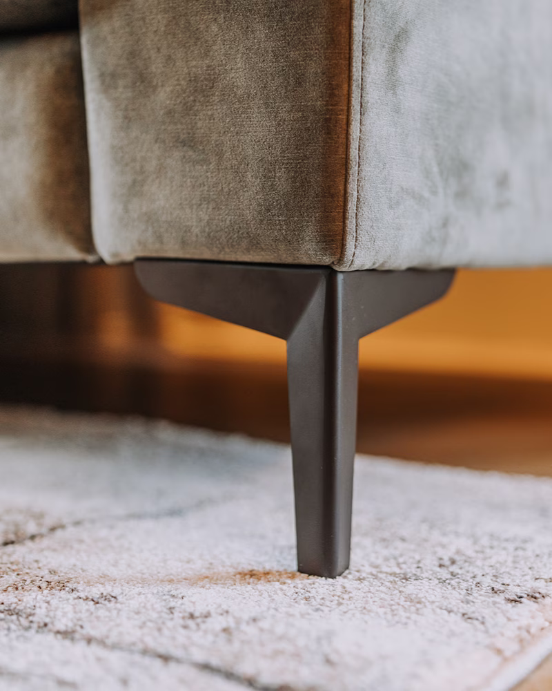 Een close-up van een moderne bankpoot van donker hout of metaal, met daarboven een zachte grijze stoffen bekleding en daaronder een lichtgrijs tapijt met textuur — de perfecte inspiratiebron als je een bank wilt uitzoeken. De achtergrond is zacht vervaagd in warme tinten.