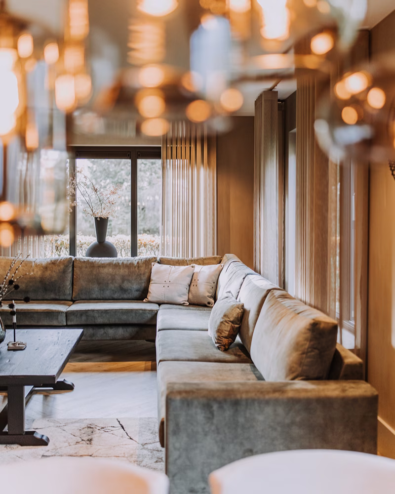 Een gezellige woonkamer met een grote grijze L-vormige bank – perfect voor wie op zoek is naar een bank – neutrale kussens, een houten salontafel, zacht natuurlijk licht dat door grote ramen naar binnen valt, beige gordijnen en wazige, warme hanglampen op de voorgrond.